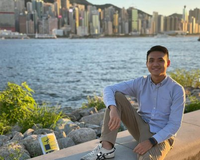 Alex Pak smiling while sitting on a waterfront wall with a city skyline behind him
