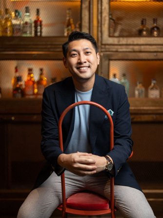 Portrait of Keith Ng, smiling in a navy blazer and seated on a red chair