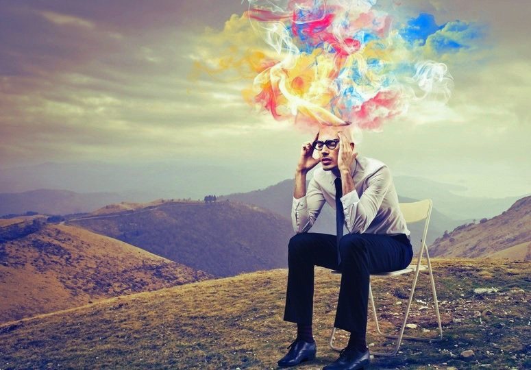 A man in glasses sitting on a folding chair on a hillside, with colourful smoke rising from his head