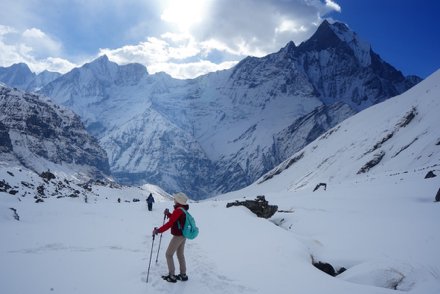 A hiker in a red jacket walking through deep snow towards a snow-covered mountain peak